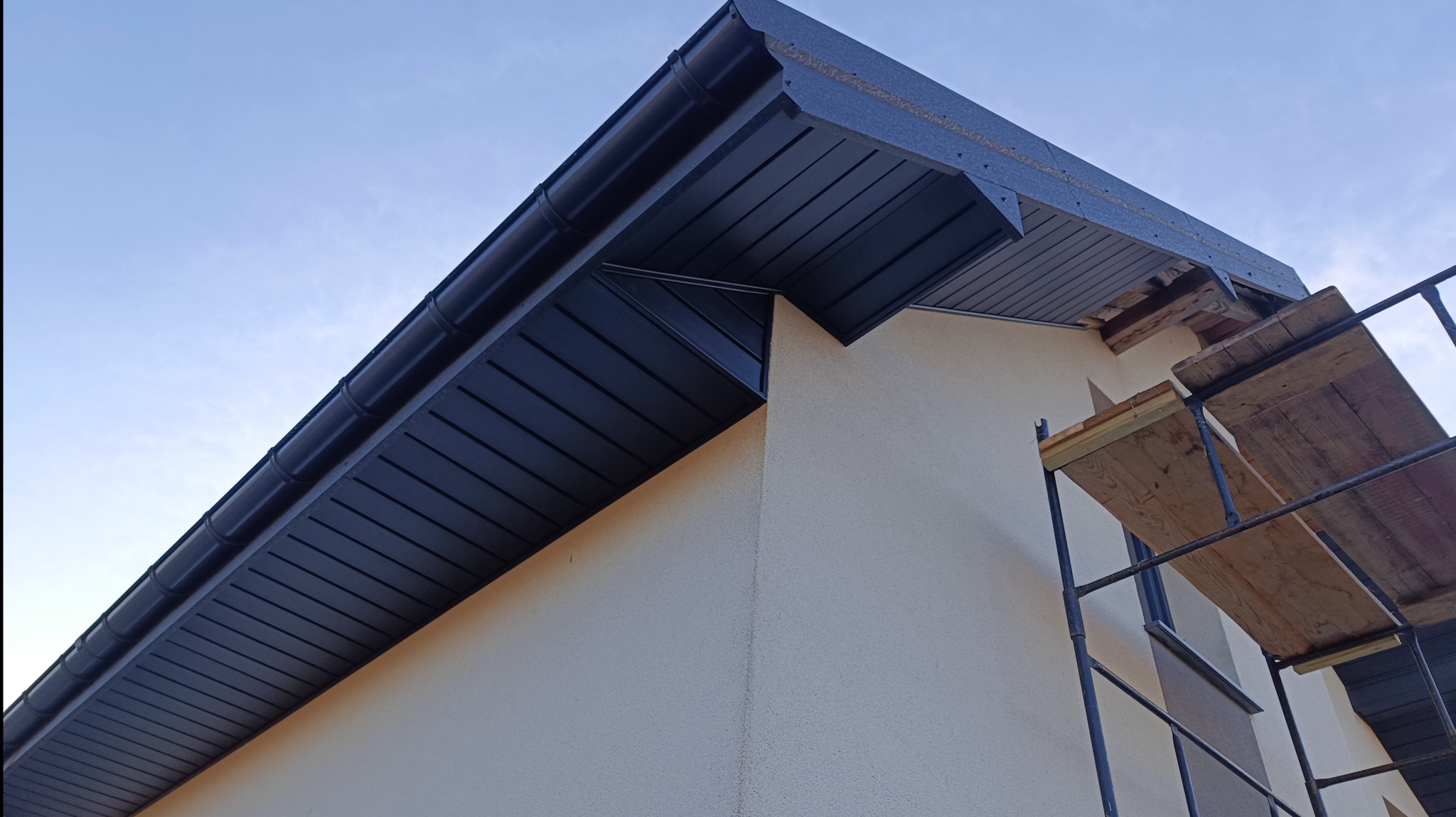 Nowoczesne, grafitowe podbitki dachowe z PVC, zamontowane na budynku o jasnej elewacji. Widoczne rusztowanie. Ujęcie z dołu, podkreślające estetykę wykończenia.