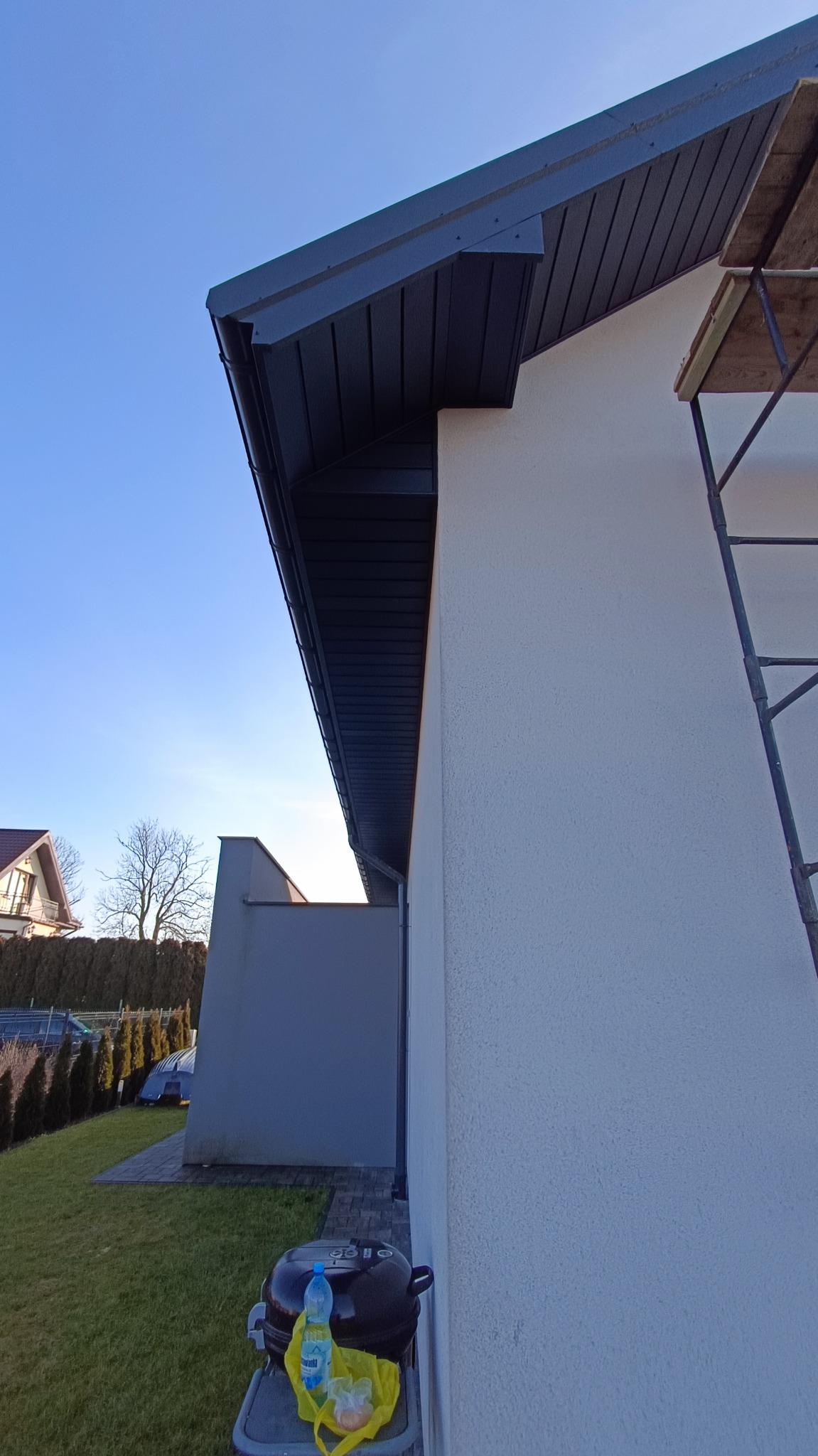 Detale wykończenia elewacji domu z ciemną podbitką dachową i fragmentem rusztowania. Widoczne elementy ogrodu i sąsiedni budynek w tle.