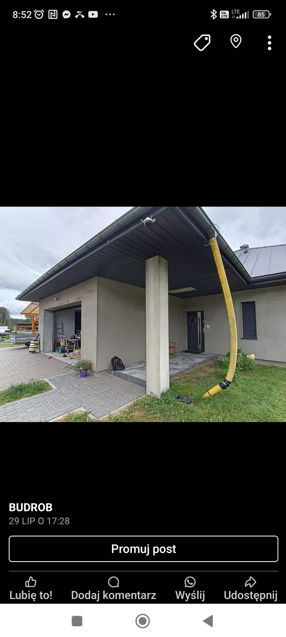 Nowoczesny dom jednorodzinny w trakcie wykończenia, z widoczną elewacją w jasnych odcieniach szarości i żółtą rurą spustową. Widoczny fragment kostki brukowej.