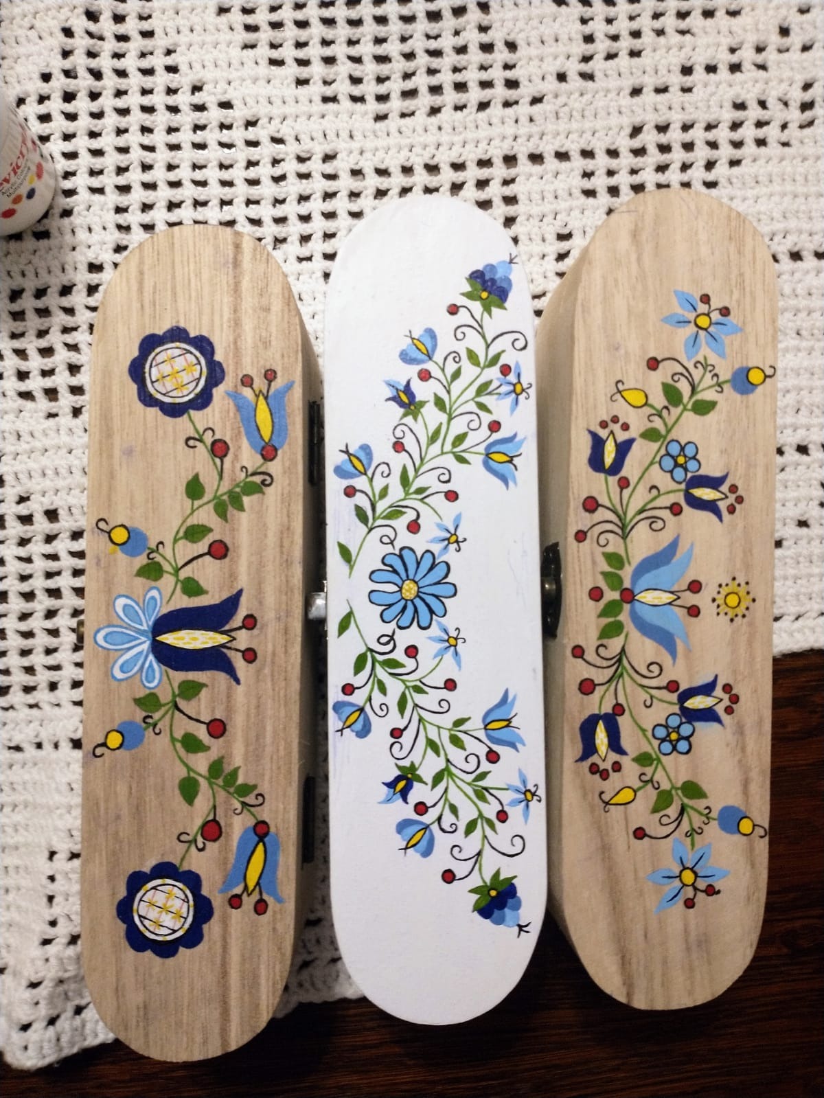 Ręcznie malowane, drewniane pudełka z motywami kwiatowymi w stylu ludowym, ustawione na białej, ażurowej serwecie.