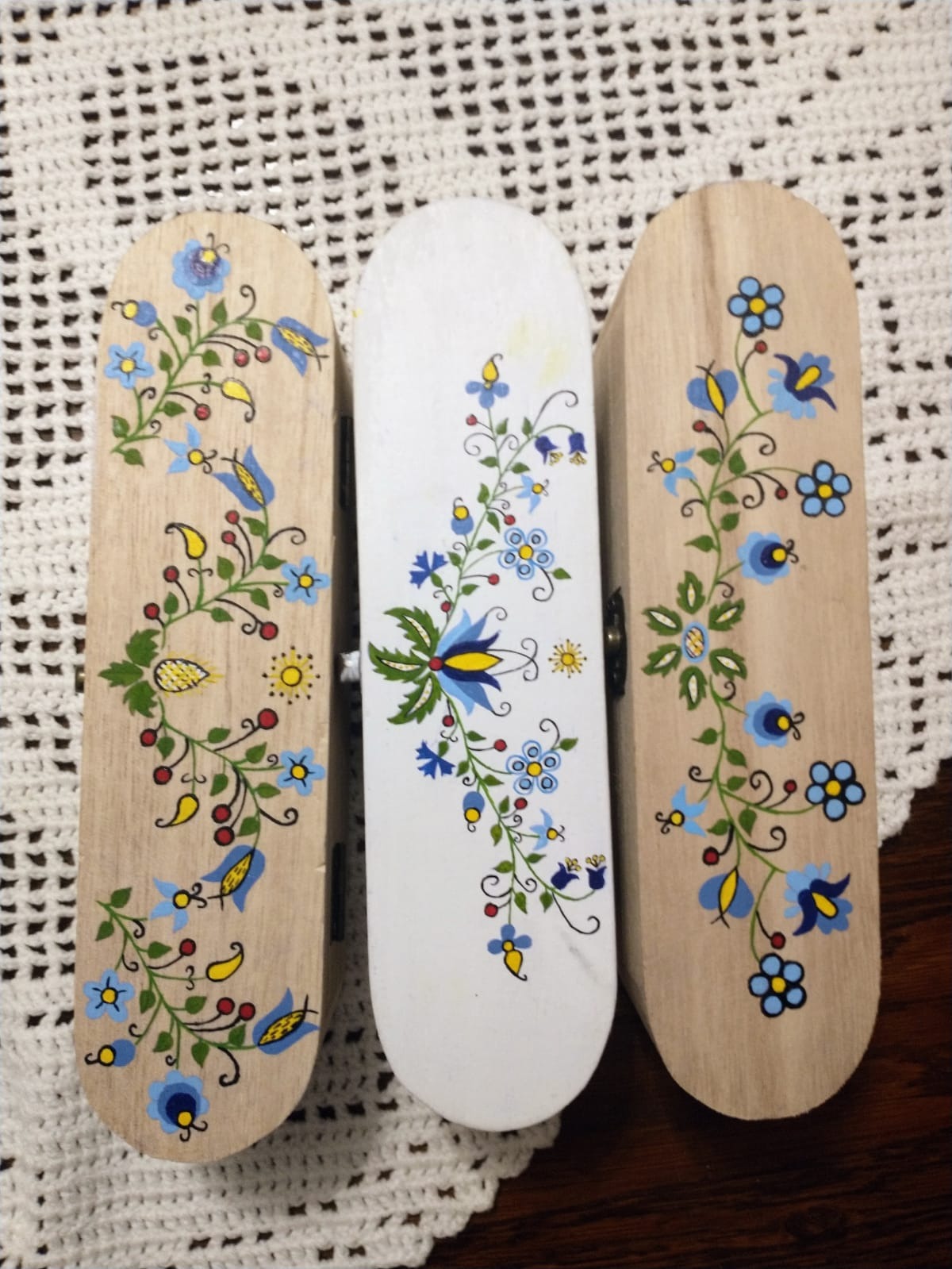 Trzy drewniane, ręcznie zdobione pudełka z motywami kwiatowymi w stylu ludowym, ułożone na białej serwetce.