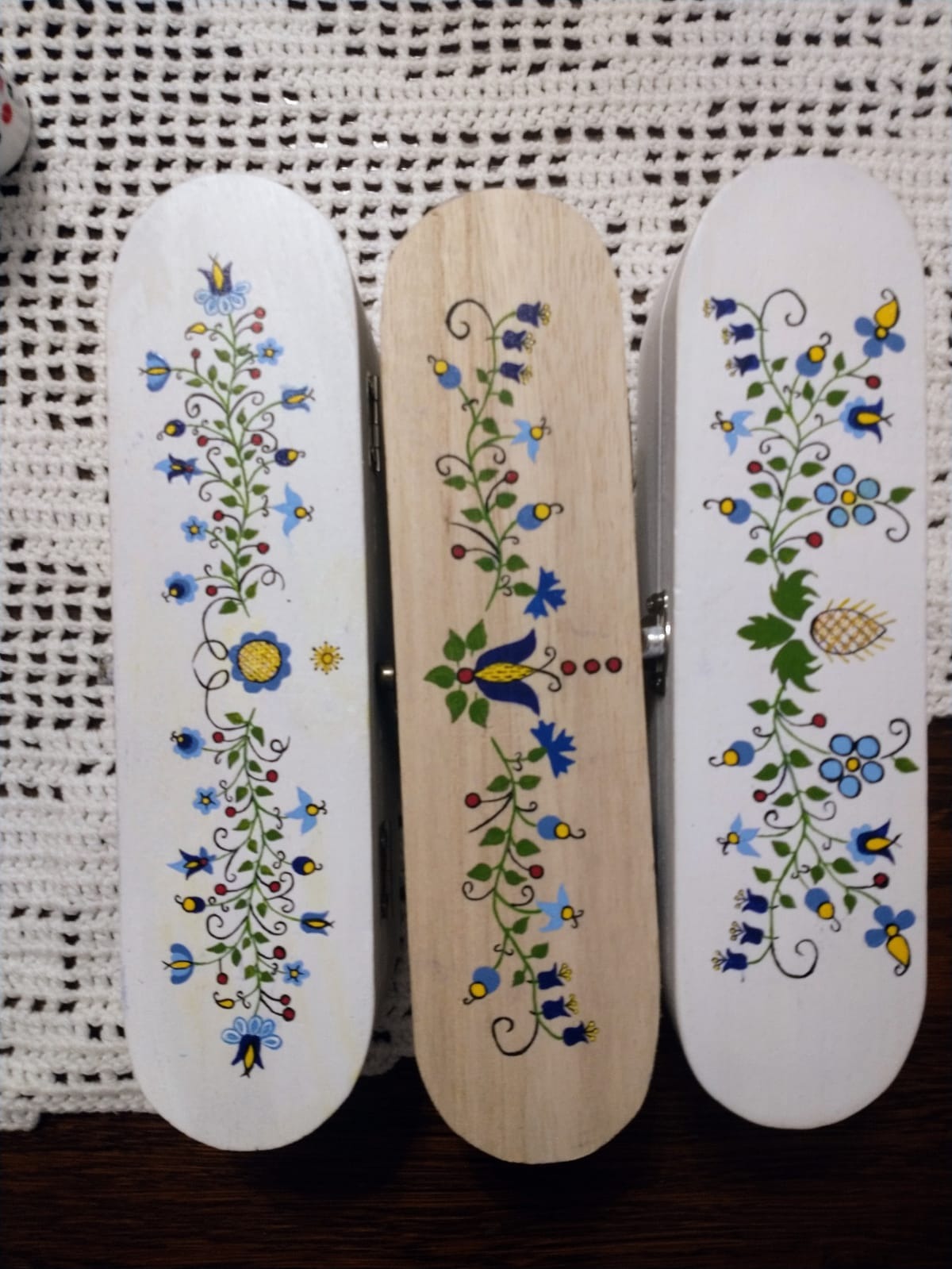 Trzy ręcznie malowane, drewniane pudełka z motywami kwiatowymi w stylu ludowym, ułożone na białej, ażurowej serwecie.