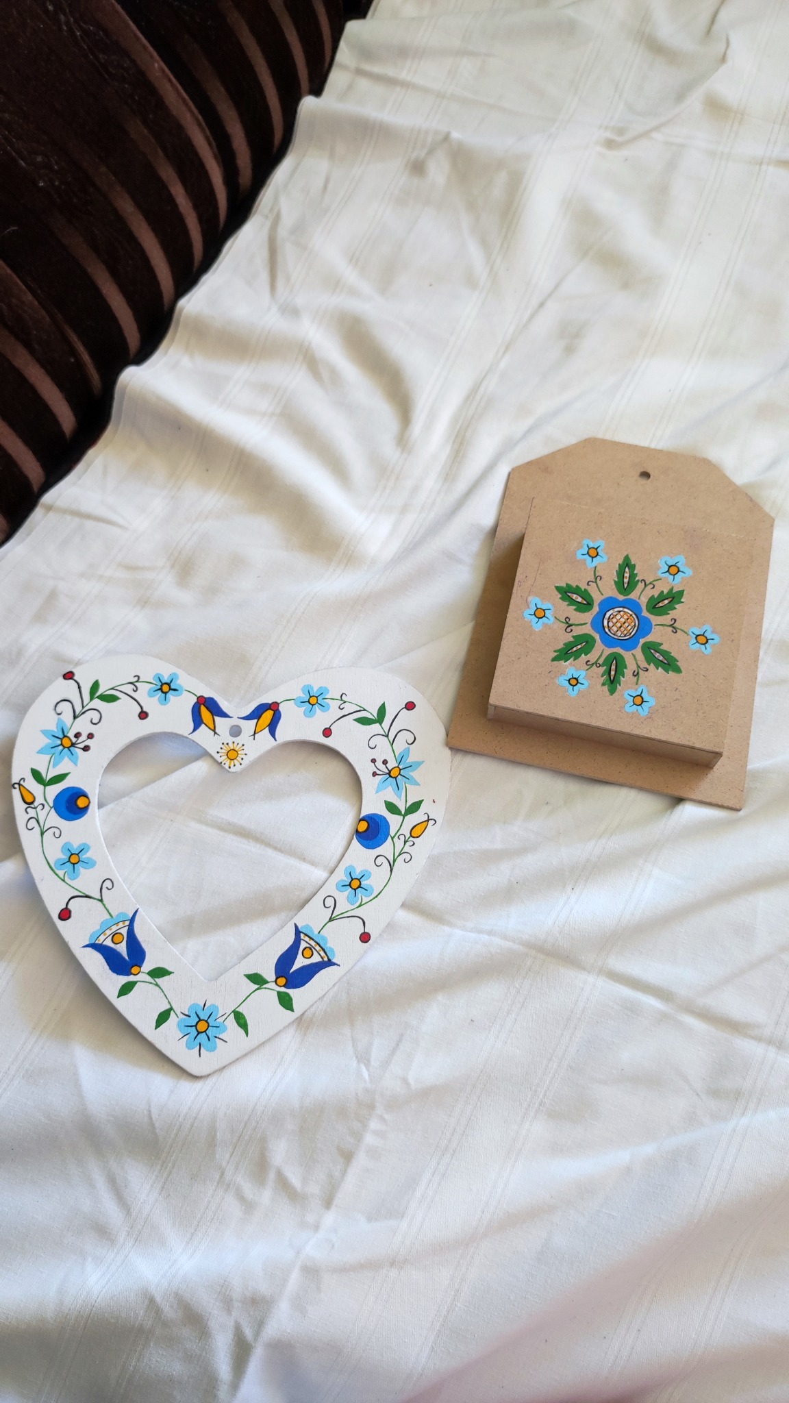 Ręcznie malowana drewniana ramka w kształcie serca z motywami kwiatowymi i ptasimi, leżąca obok malowanej drewnianej kartki na białym, pogniecionym tle.