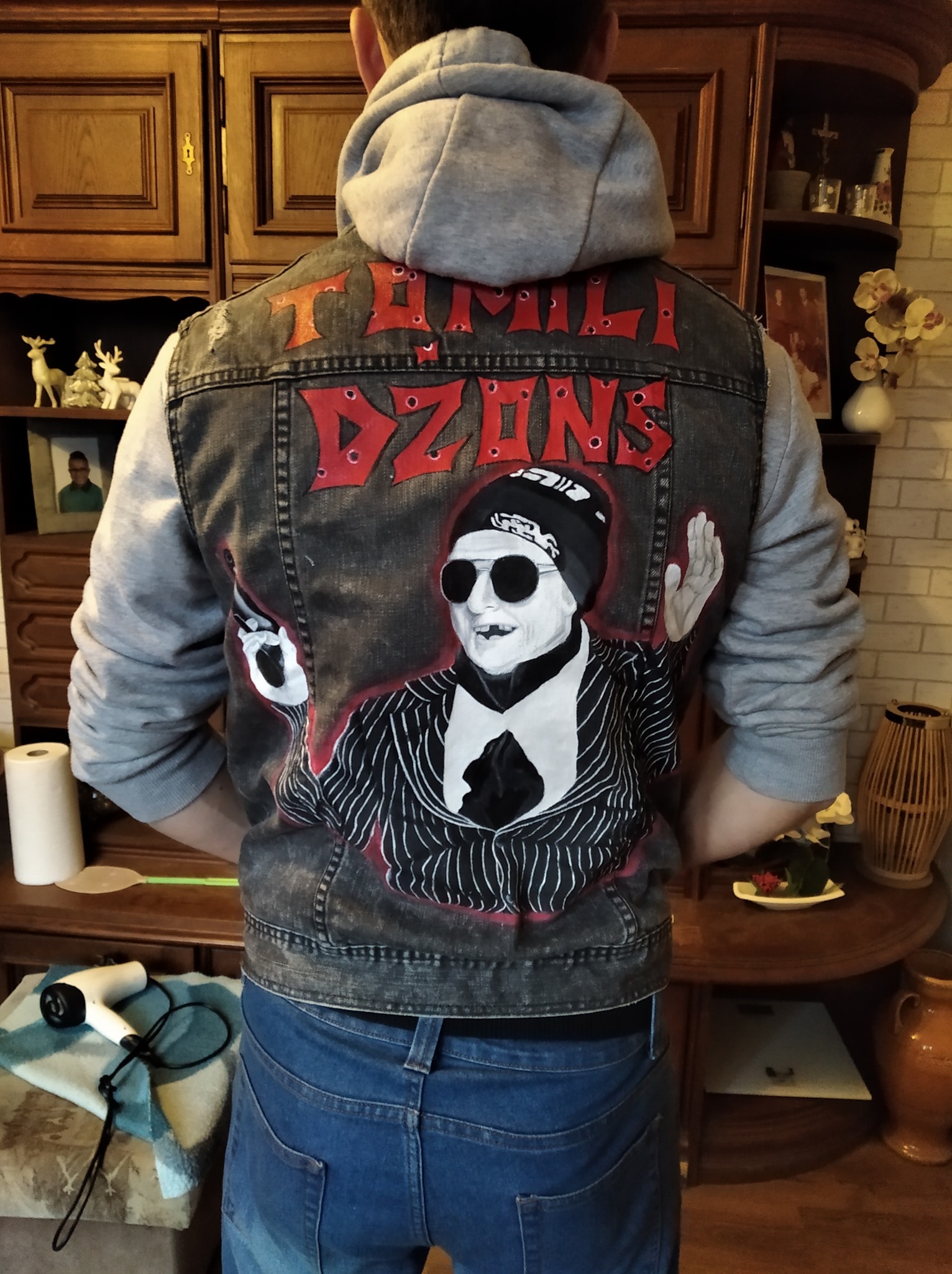 Osoba w dżinsowej kurtce z ręcznie malowanym portretem w stylu gangstera i napisem 'FAMILY DZONS' na plecach, stojąca tyłem na tle drewnianej komody i okrągłego stolika.
