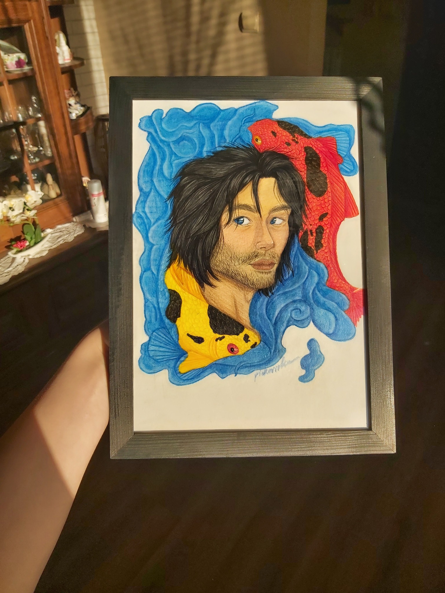 Ręcznie rysowany portret mężczyzny z rybami Koi w niebieskiej wodnej scenerii, oprawiony w ramkę, trzymany w dłoni w słonecznym świetle.