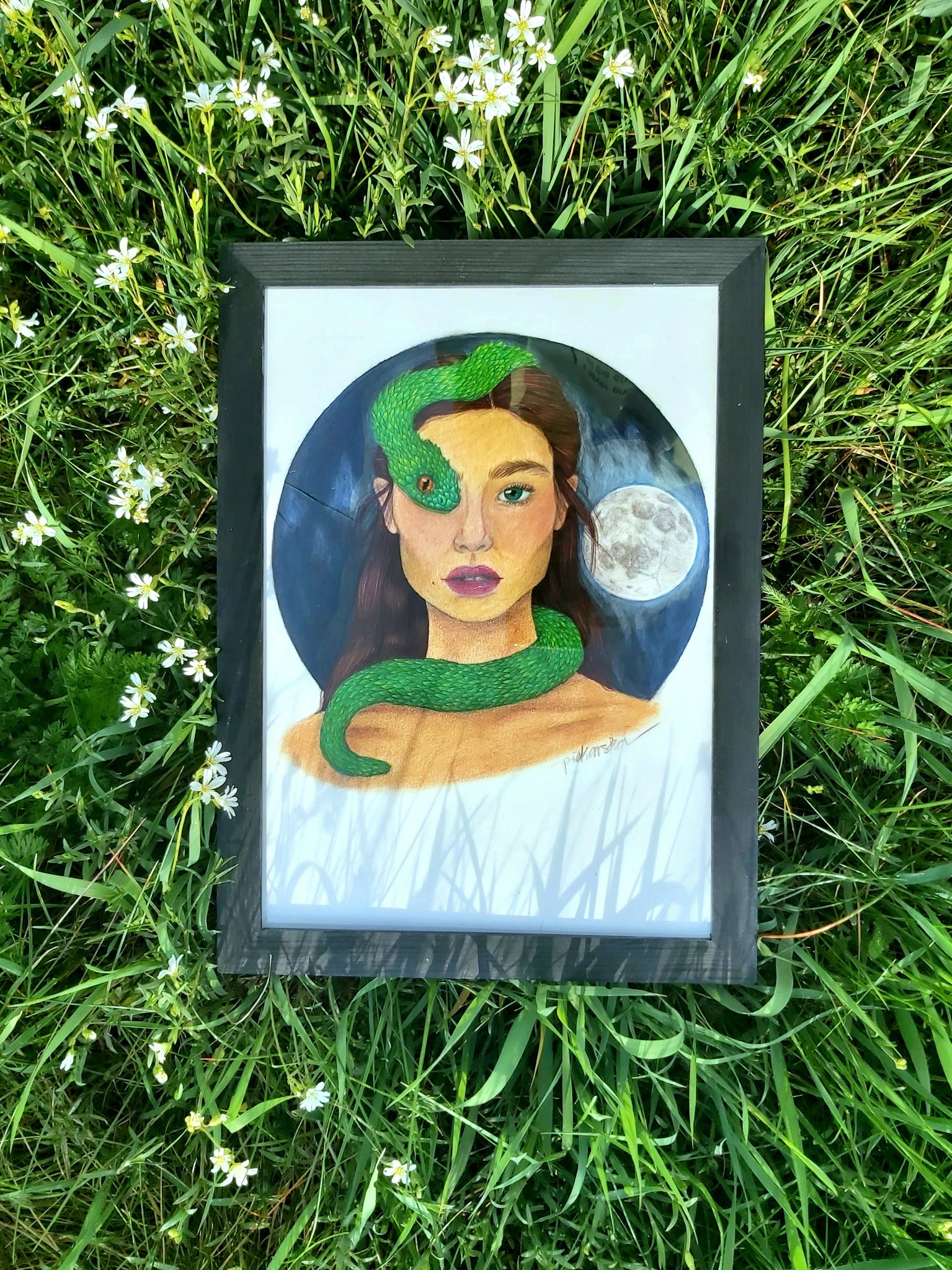 Oprawiony portret kobiety z zielonym wężem owiniętym wokół głowy, leżący na trawie z białymi kwiatami, z widocznym podpisem artysty w prawym dolnym rogu.