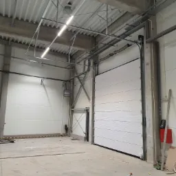 Częściowo otwarta, biała segmentowa brama garażowa w surowym, industrialnym wnętrzu hali produkcyjnej, widoczne elementy konstrukcyjne i instalacyjne pod sufitem.