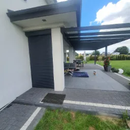 Zewnętrzna roleta w kolorze antracytowym, zamontowana na nowoczesnym domu z tarasem i pergolą, widok z boku, trawnik z dekoracyjnymi obrzeżami.