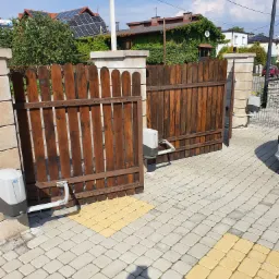 Otwarta, dwuskrzydłowa brama wjazdowa z brązowego drewna, z automatyką DEA, osadzona między kamiennymi słupkami, na podjeździe z szarej kostki brukowej z żółtymi akcentami.