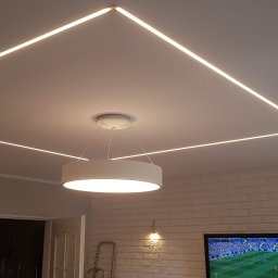 RemoGresik - Nowoczesne oświetlenie sufitowe LED w kształcie kwadratu, z centralnie umieszczoną okrągłą lampą wiszącą, na tle ściany z białej cegły i włączonego telewizora.