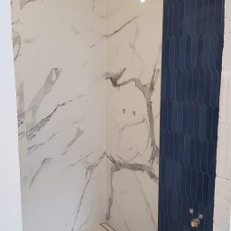 RemoGresik - Wnętrze łazienki w trakcie wykańczania: białe płytki imitujące marmur na ścianach i podłodze, pionowy pas ciemnoniebieskich płytek o podłużnym kształcie, widoczny odpływ liniowy w podłodze, surowe...