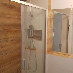 RemoGresik - Wykończona kabina prysznicowa z przesuwnymi drzwiami, obudowana płytkami imitującymi drewno, widok z lustra z fragmentem instalacji elektrycznej