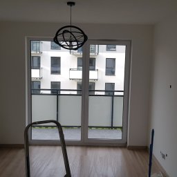 RemoGresik - Pusty pokój w trakcie prac wykończeniowych, z widoczną drabiną, grzejnikiem i lampą sufitową w stylu industrialnym; ściany świeżo pomalowane na biało, widok z okna na budynek z balkonami.