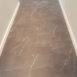 RemoGresik - Szare płytki podłogowe z marmurkowym wzorem ułożone w korytarzu z białą listwą przypodłogową i drzwiami w tle.