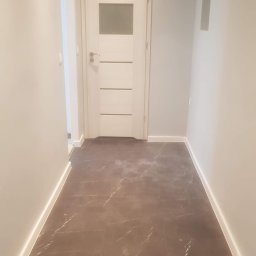 RemoGresik - Wykończony korytarz z brązowymi płytkami imitującymi marmur, białe ściany i drzwi z przeszkleniem, widoczny fragment instalacji na suficie.