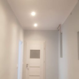 RemoGresik - Korytarz po odświeżeniu: szare ściany, białe drzwi z przeszkleniem, ciemna podłoga, trzy punktowe lampy sufitowe.