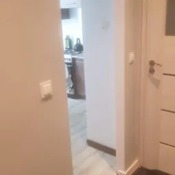 Widok z korytarza na kuchnię z fragmentem podłogi przed ułożeniem paneli, widoczne listwy przypodłogowe i włącznik światła.