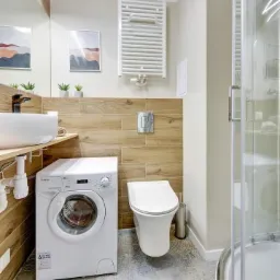 Nowoczesna łazienka z drewnopodobnymi płytkami, białą umywalką nablatową, pralką i kabiną prysznicową z chromowanymi detalami.