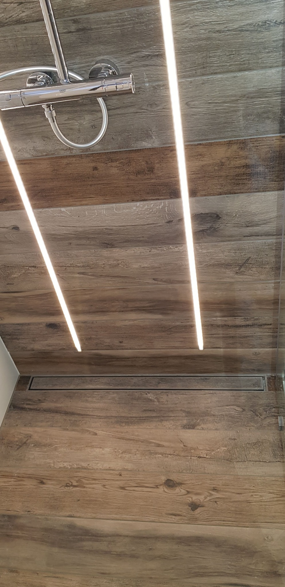 Kątowe ujęcie ściany prysznica wyłożonej płytkami imitującymi drewno, z widocznym odpływem liniowym i oświetleniem LED oraz chromowaną armaturą Grohe.