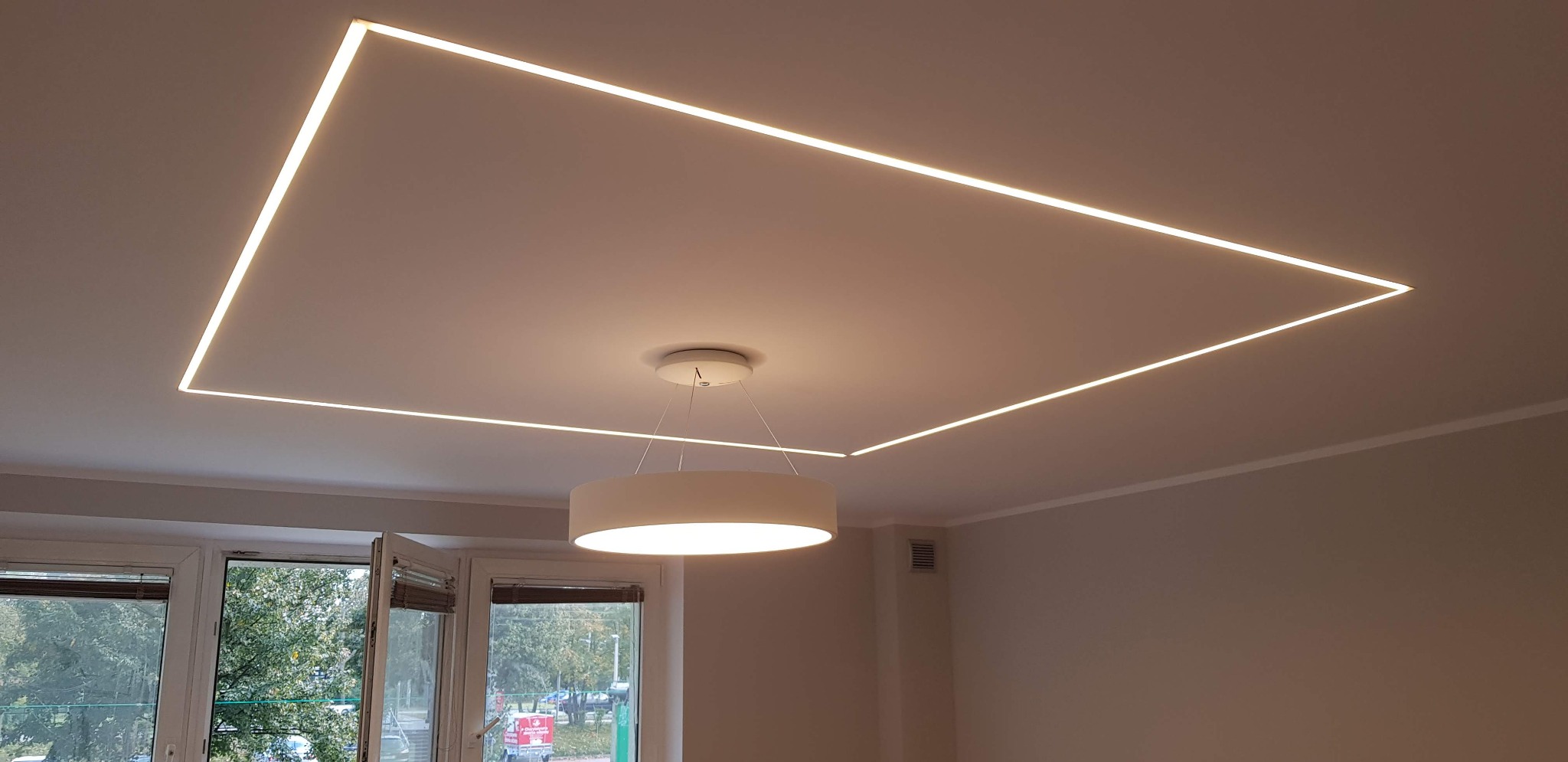 Nowoczesne oświetlenie sufitowe: prostokątna linia LED wpuszczona w sufit, uzupełniona wiszącą, okrągłą lampą z abażurem, widok z dołu na jasne wnętrze z oknem.