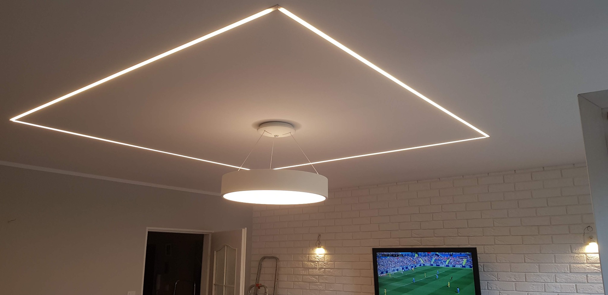 Nowoczesne oświetlenie sufitowe LED w kształcie kwadratu, z centralnie umieszczoną okrągłą lampą wiszącą, na tle ściany z białej cegły i włączonego telewizora.
