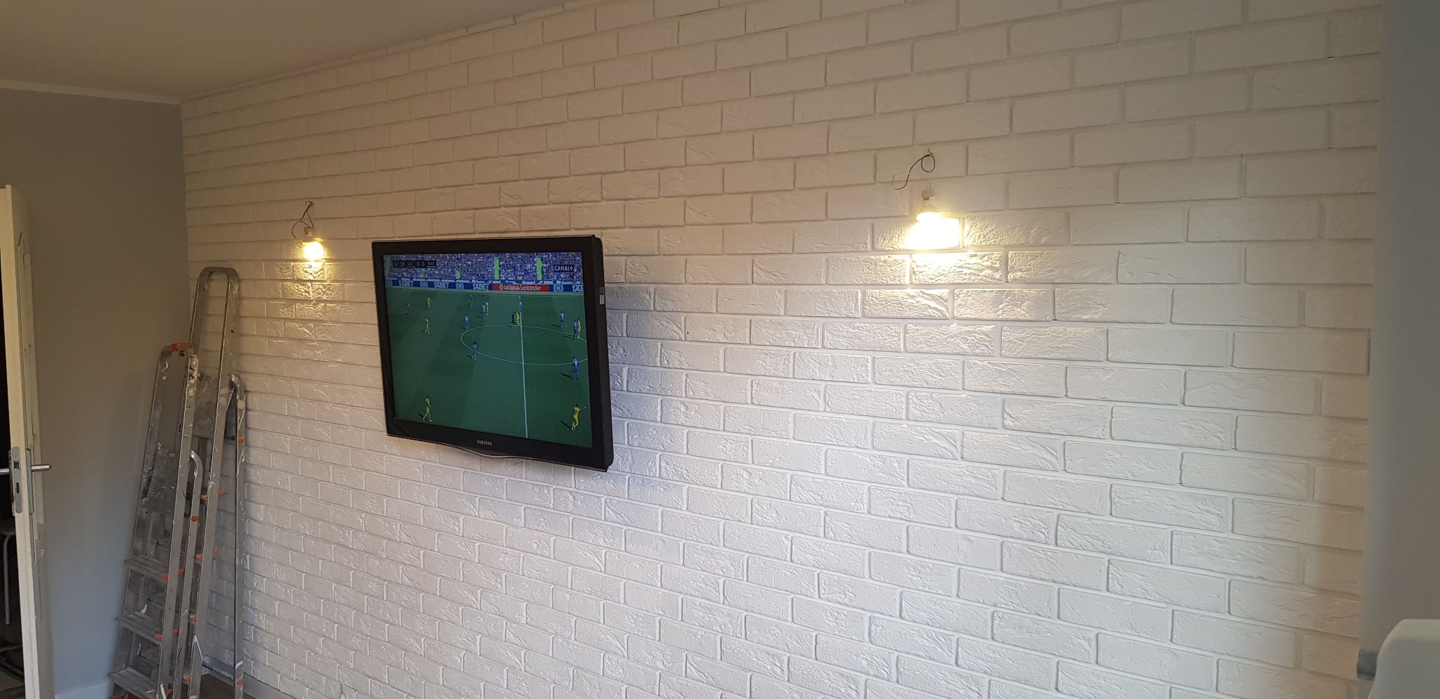 Ściana wyłożona białą cegłą dekoracyjną z zamontowanym telewizorem, oświetlona kinkietami, z drabiną opartą o ścianę w rogu pokoju.