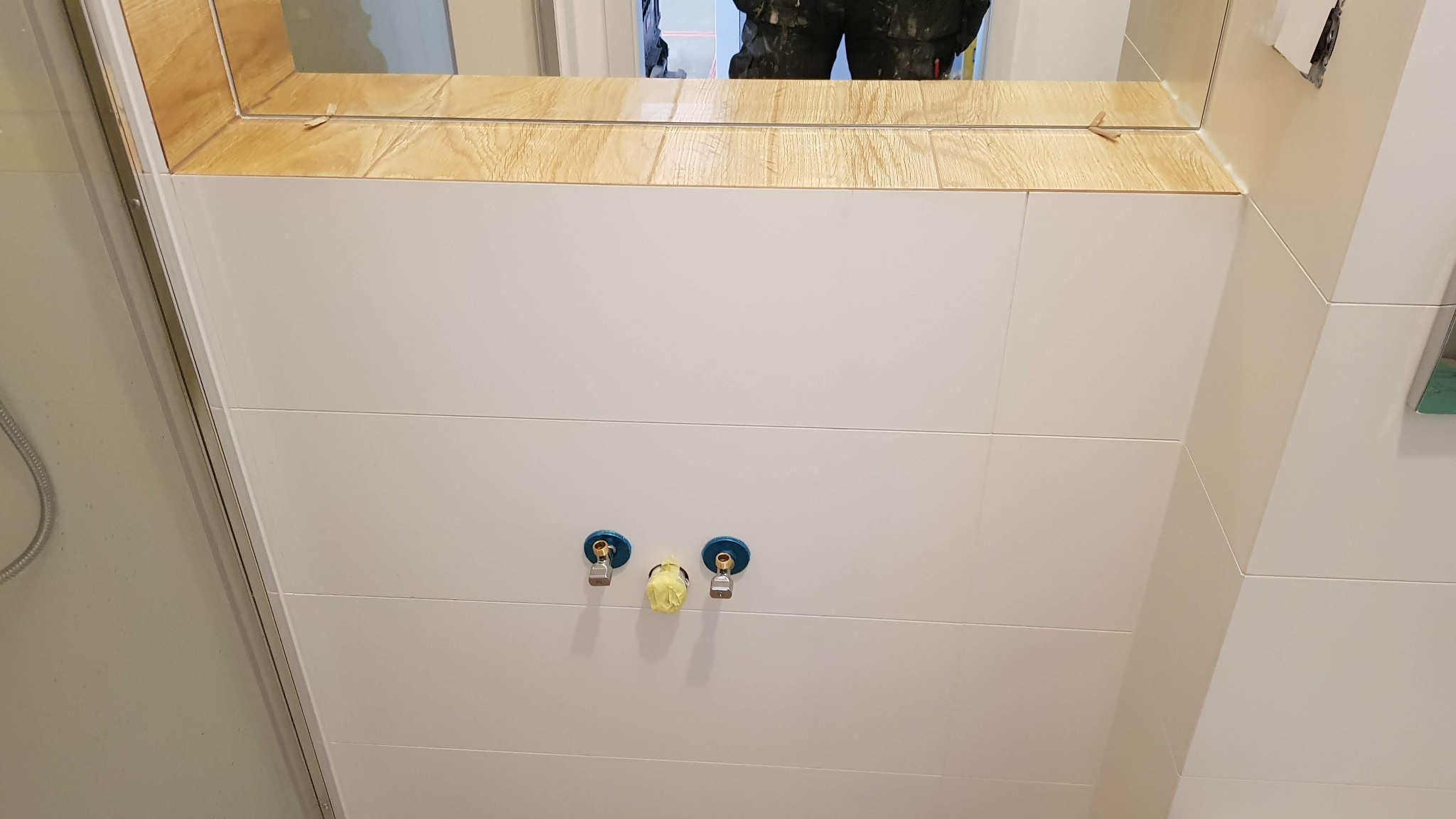 Ściana łazienki z białymi, poziomymi płytkami, trzema wystającymi zaworami zakrytymi taśmą, lustrem z drewnopodobną obudową u góry i fragmentem kabiny prysznicowej po lewej stronie.