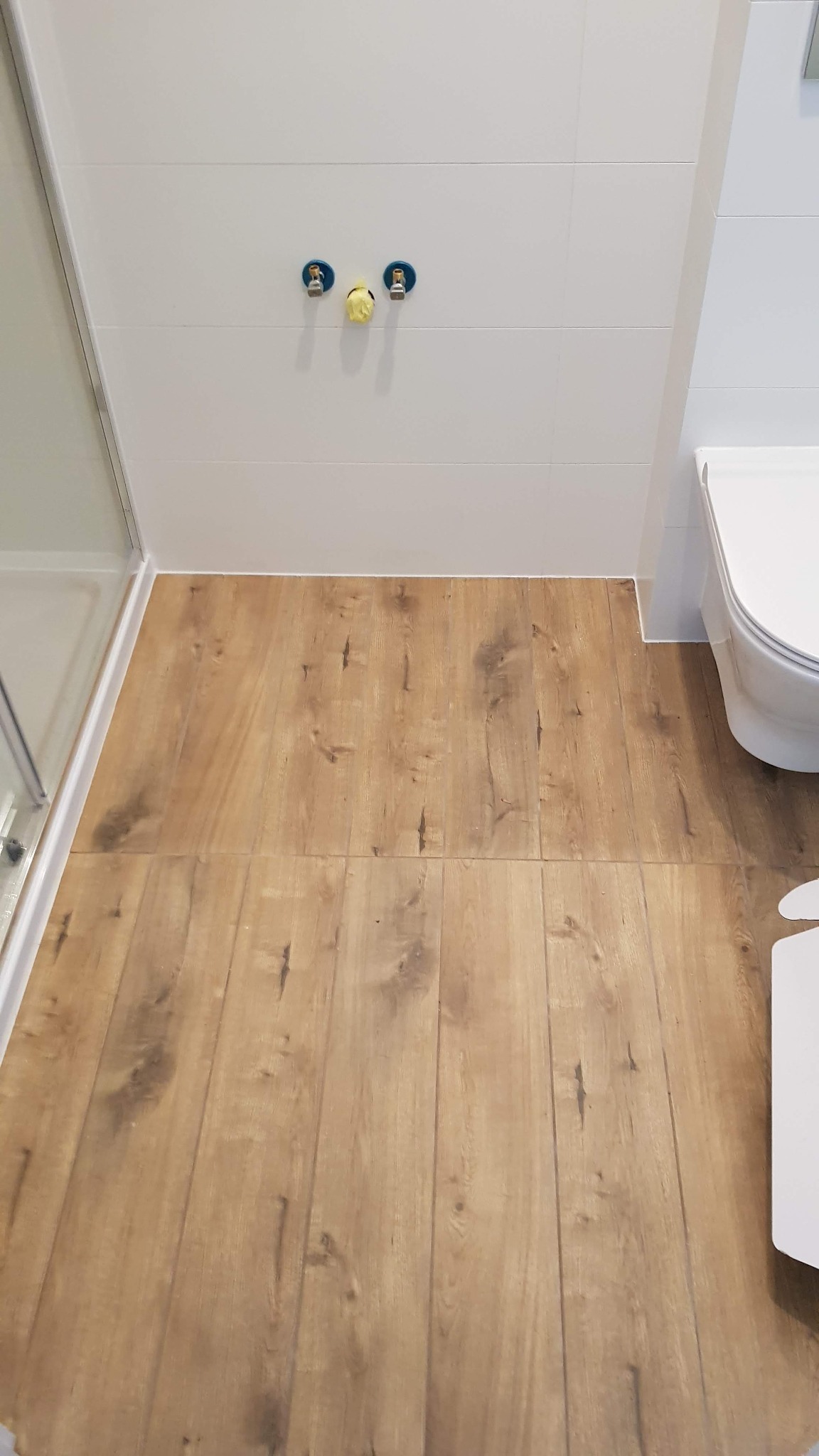 Wykończona łazienka z panelami podłogowymi imitującymi drewno, białe płytki ścienne, widoczne przyłącza wodne z zaślepkami i fragment toalety.