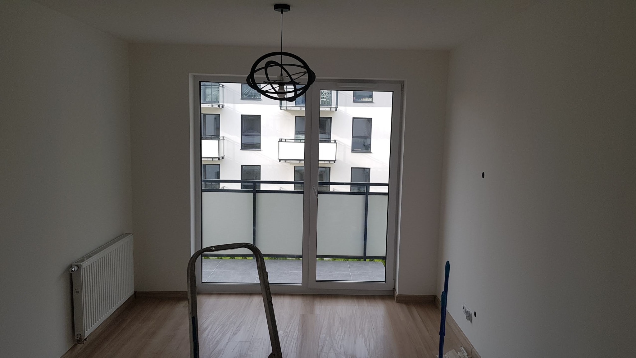 Pusty pokój w trakcie prac wykończeniowych, z widoczną drabiną, grzejnikiem i lampą sufitową w stylu industrialnym; ściany świeżo pomalowane na biało, widok z okna na budynek z balkonami.
