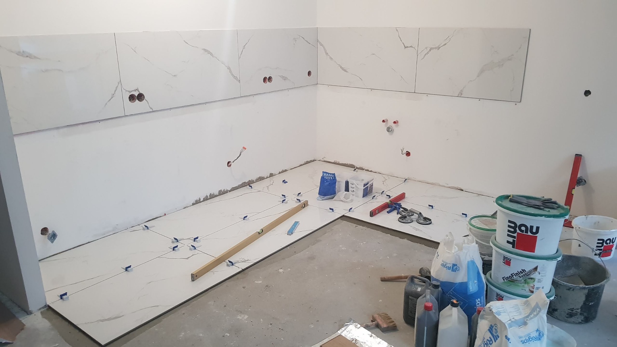 Wnętrze kuchni w trakcie prac wykończeniowych: układanie białych płytek imitujących marmur na podłodze i ścianach, widoczne krzyżyki dystansowe, poziomica, puszki elektryczne, wiadro z klejem...