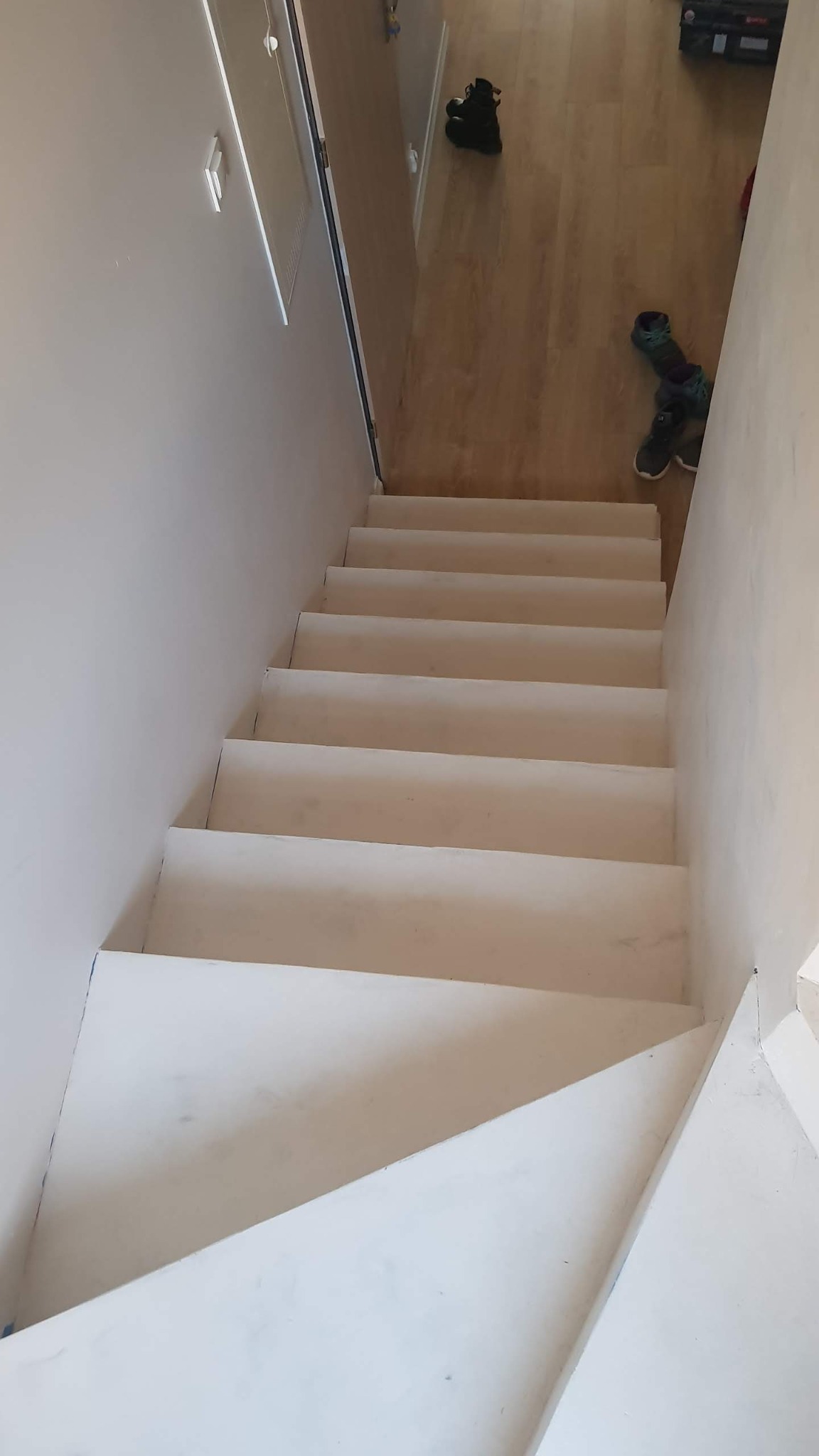 Widok z góry na białe, pomalowane schody wewnątrz domu, z widocznymi butami i skrzynką narzędziową na podłodze na górze schodów. Ściany pomalowane na jasny kolor, widoczne niedoskonałości malowania.