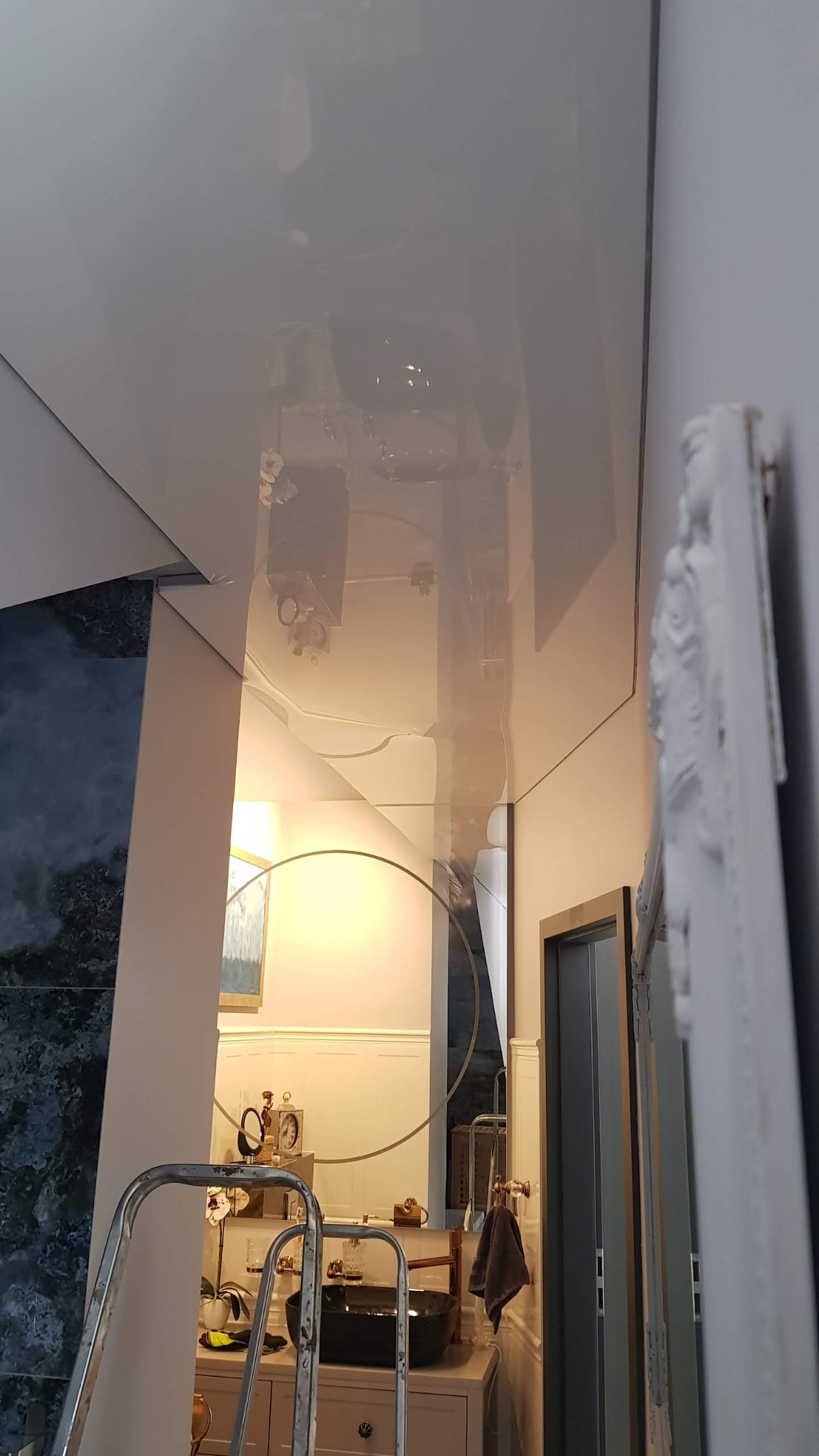 Błyszczący, biały sufit napinany w łazience, widok z dołu, częściowo zasłonięty przez drabinę i ramę lustra, z odbiciem wyposażenia łazienki.