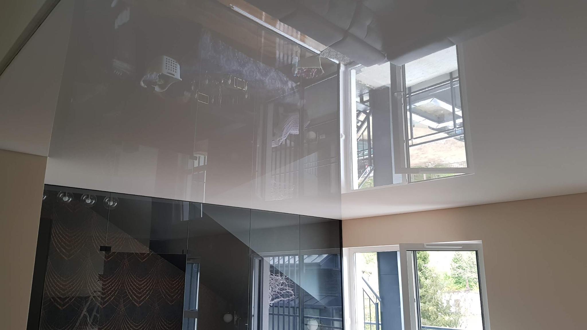 Błyszczący, szary sufit napinany z widocznym odbiciem wnętrza, okna z widokiem na zewnątrz, ściany w odcieniach beżu, nowoczesne oświetlenie punktowe.