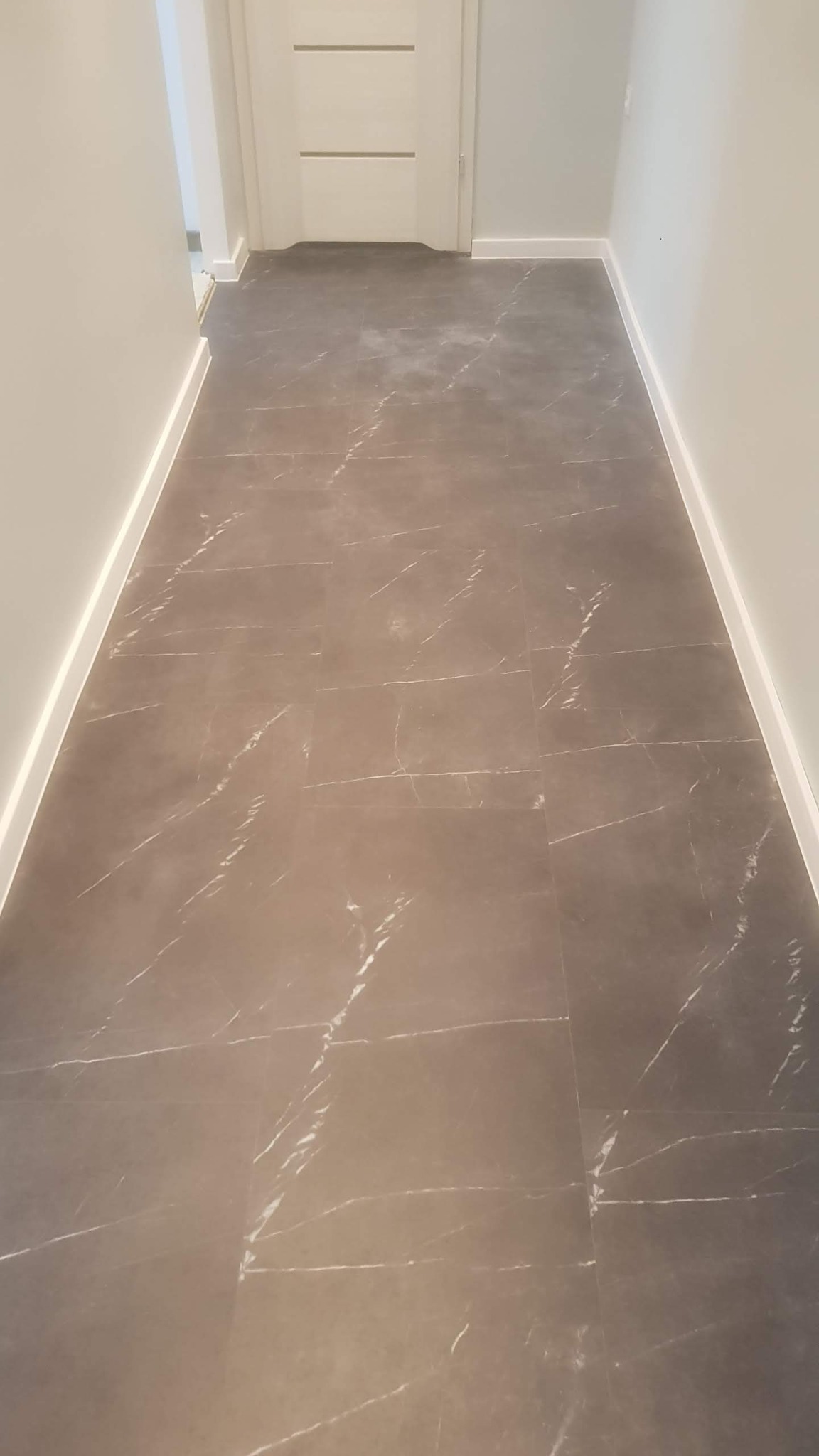 Szare płytki podłogowe z marmurkowym wzorem ułożone w korytarzu z białą listwą przypodłogową i drzwiami w tle.