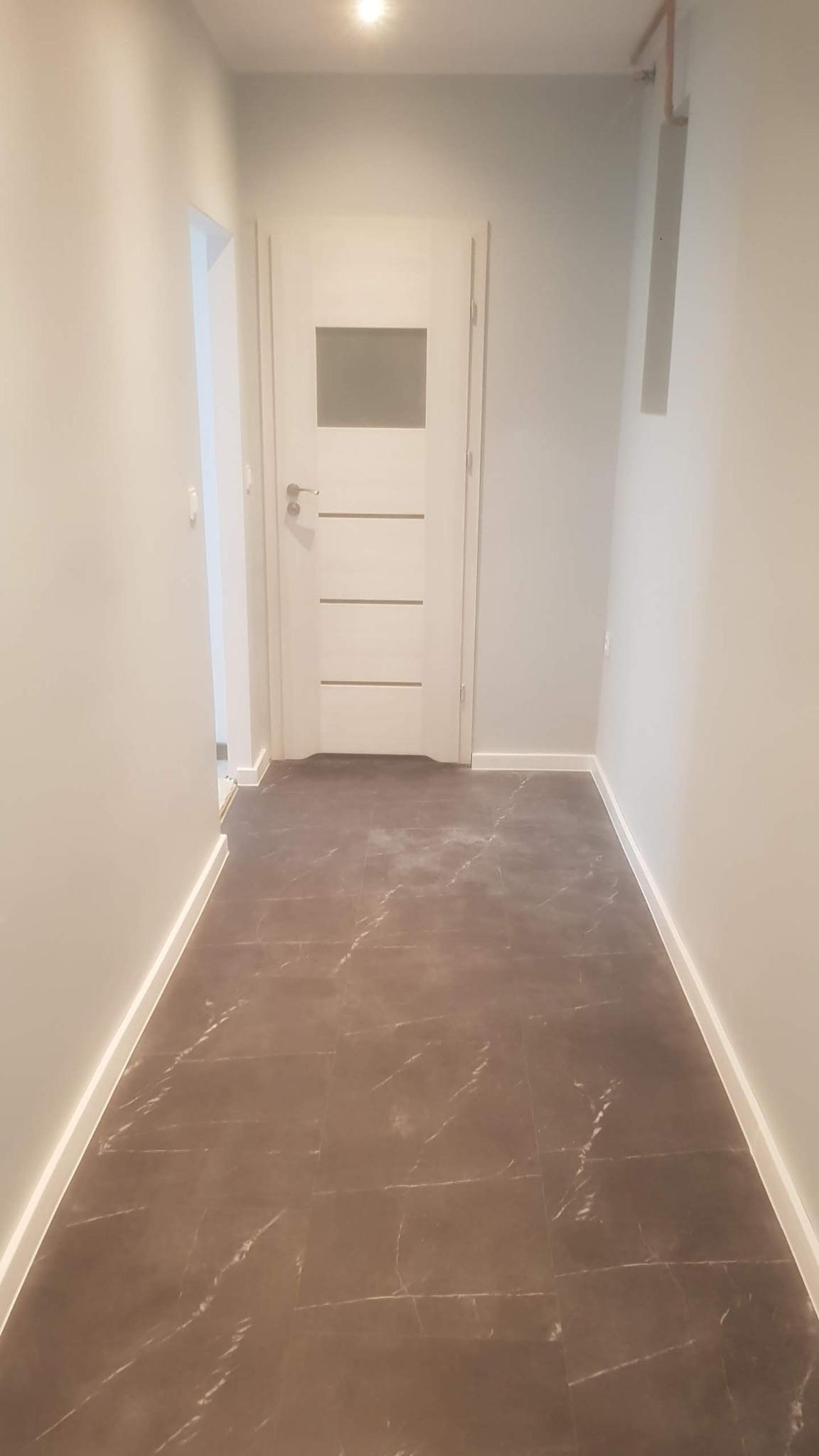 Wykończony korytarz z brązowymi płytkami imitującymi marmur, białe ściany i drzwi z przeszkleniem, widoczny fragment instalacji na suficie.