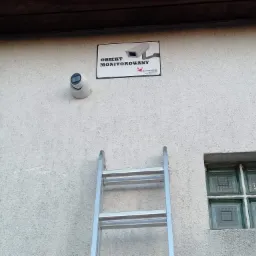Kamera monitoringu zamontowana na elewacji budynku obok tabliczki informującej o monitoringu, drabina oparta o ścianę, okno z luksferów w tle.