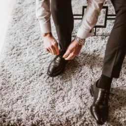 Mężczyzna w eleganckiej koszuli i spodniach zawiązuje sznurowadła w czarnych skórzanych butach na tle puszystego szarego dywanu, widoczny zegarek na nadgarstku.