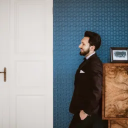 Uśmiechnięty mężczyzna w bordowym smokingu, oparty o białe drzwi na tle ściany z geometrycznym wzorem, obok drewnianego kredensu z ramką ze zdjęciem.