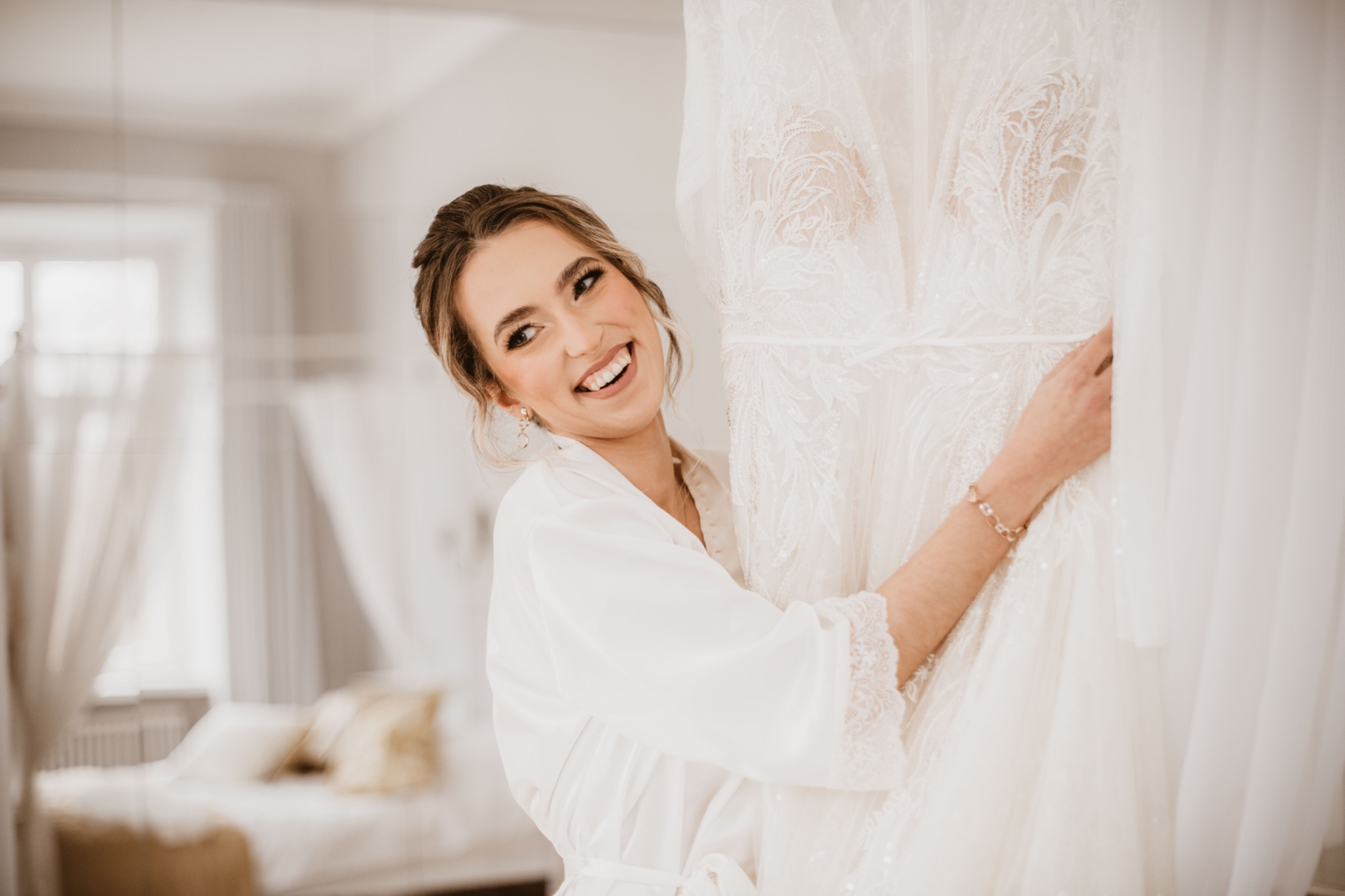 Uśmiechnięta panna młoda w białym szlafroku, stojąca obok koronkowej sukni ślubnej z delikatnymi zdobieniami, w jasnym wnętrzu sypialni z baldachimem w tle.