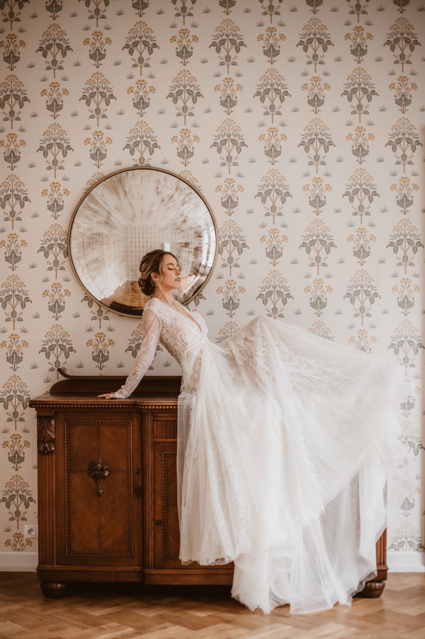 Panna młoda w koronkowej sukni z długim rękawem opiera się o antyczny kredens na tle ściany w kwiecistej tapecie i okrągłego lustra.