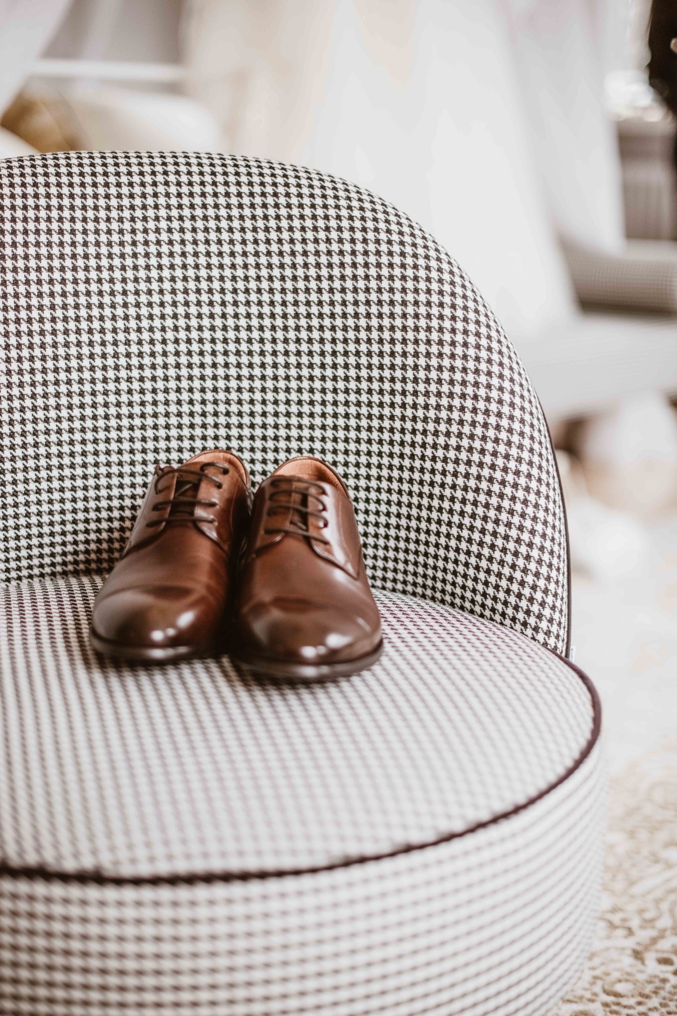 Brązowe, skórzane buty męskie ze sznurowadłami leżą na krześle obitym tkaniną w pepitkę. Delikatne światło dzienne. W tle rozmyte elementy garderoby.