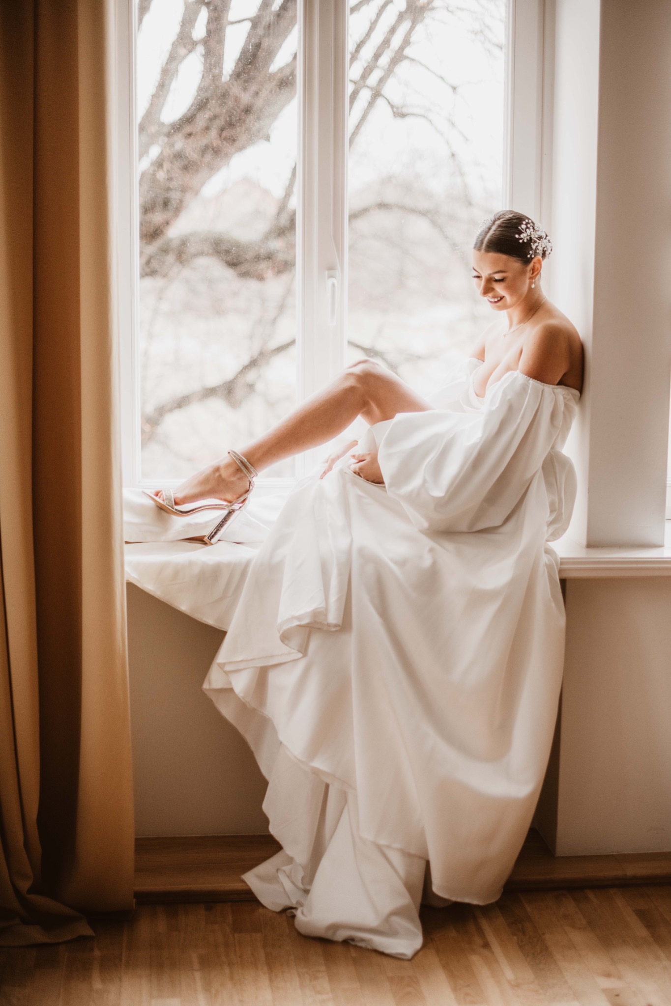 Uśmiechnięta panna młoda w białej sukni siedzi na parapecie okna, eksponując nogę w srebrnym bucie na obcasie, z delikatną ozdobą we włosach i widokiem na drzewa za oknem.