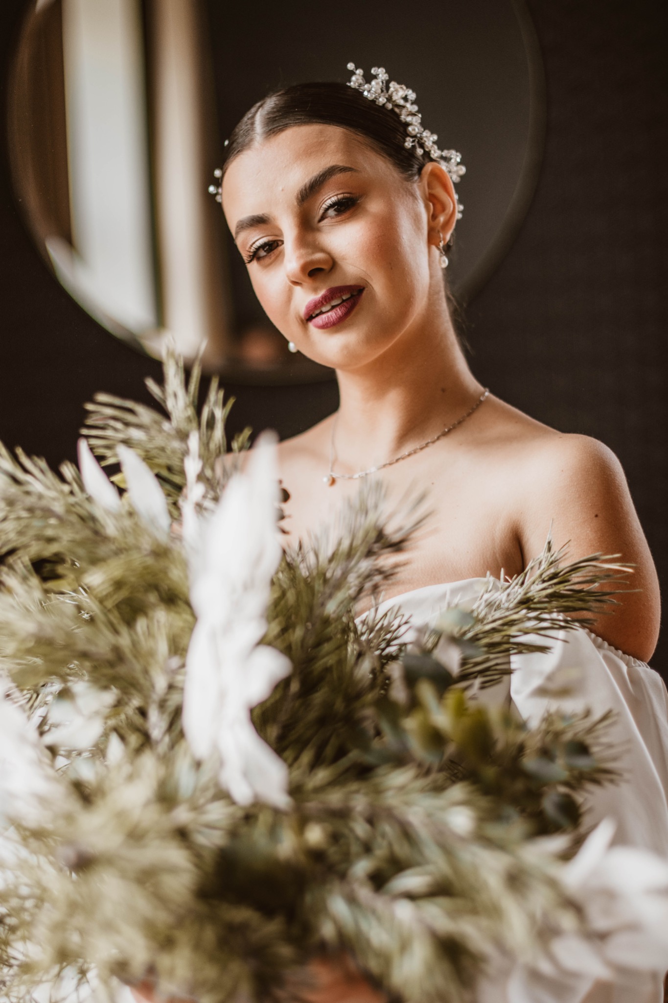 Portret panny młodej w białej sukni z bukietem z suszonych traw i kwiatów, z perłową ozdobą we włosach i delikatną biżuterią, na ciemnym tle z odbiciem okna.