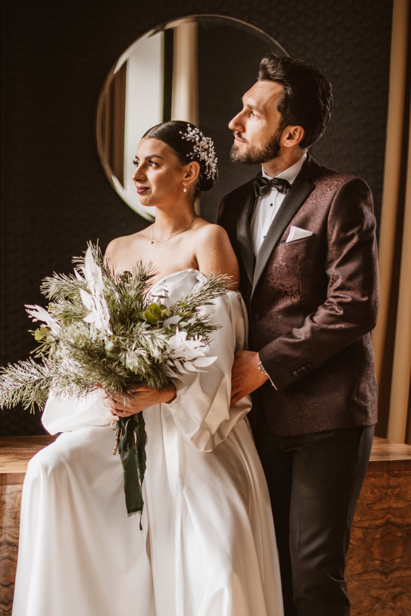 Elegancka para narzeczonych: panna młoda w białej sukni z bukietem z gałązek i pan młody w bordowym smokingu z muszką, pozują na tle ciemnej ściany z okrągłym lustrem.