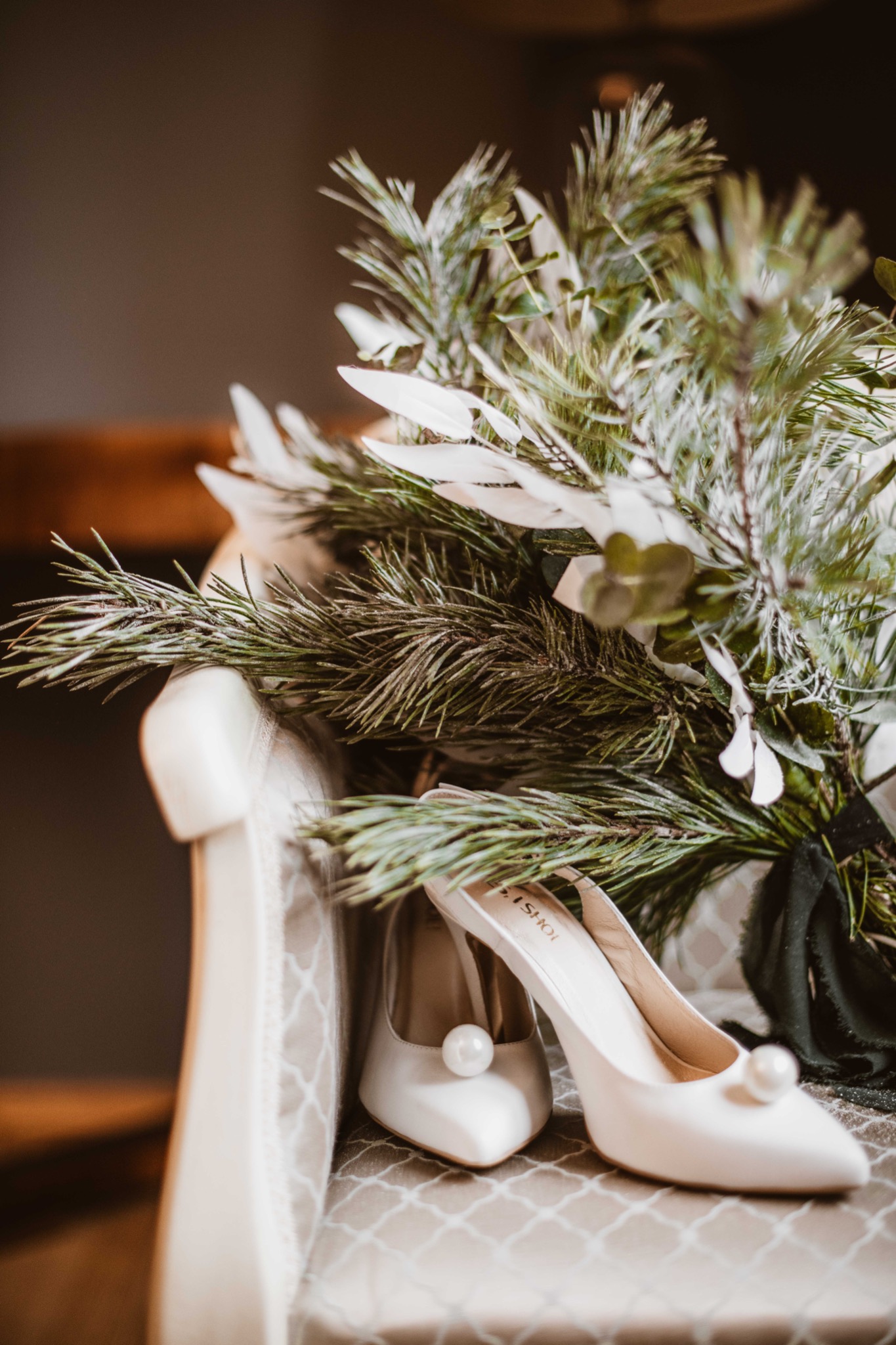 Eleganckie białe szpilki z perłami, leżące obok bukietu z gałązek iglastych i białych liści na krześle z wzorzystą tapicerką.
