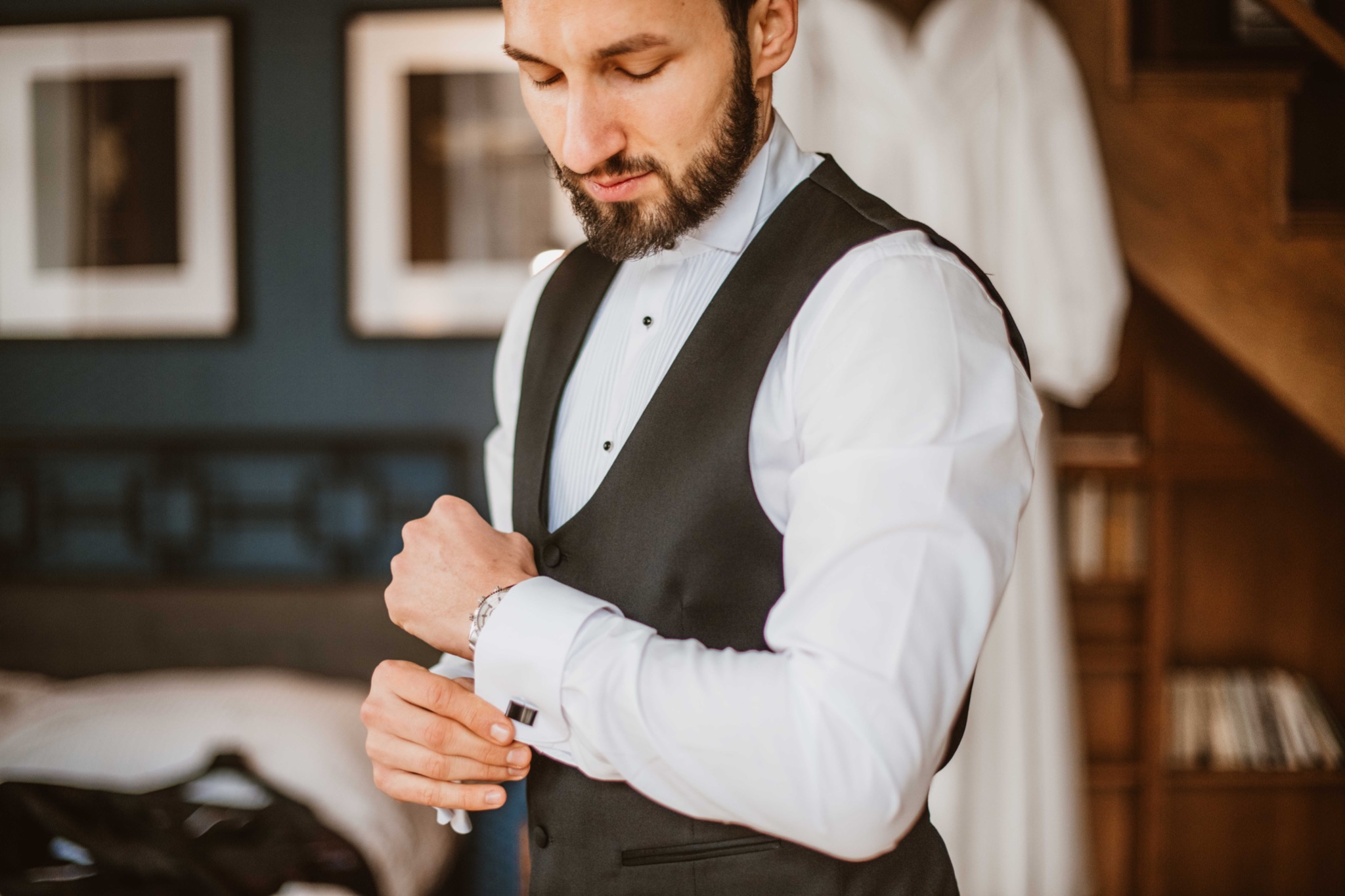Mężczyzna z brodą, w białej koszuli i czarnej kamizelce, zapinający mankiet koszuli z eleganckimi spinkami, na tle ciemnej ściany z obrazami i białej sukni ślubnej.
