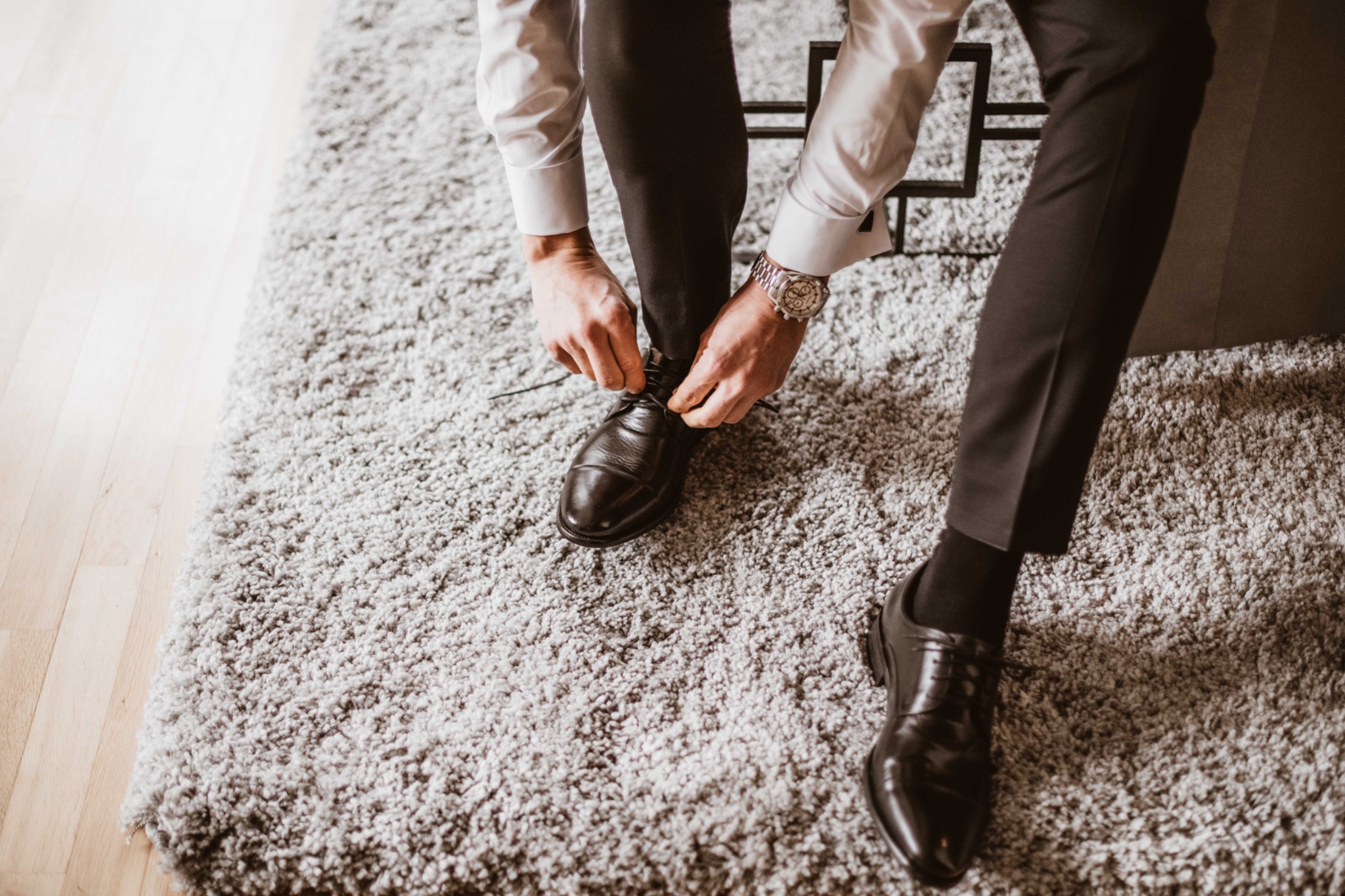 Mężczyzna w eleganckiej koszuli i spodniach zawiązuje sznurowadła w czarnych skórzanych butach na tle puszystego szarego dywanu, widoczny zegarek na nadgarstku.
