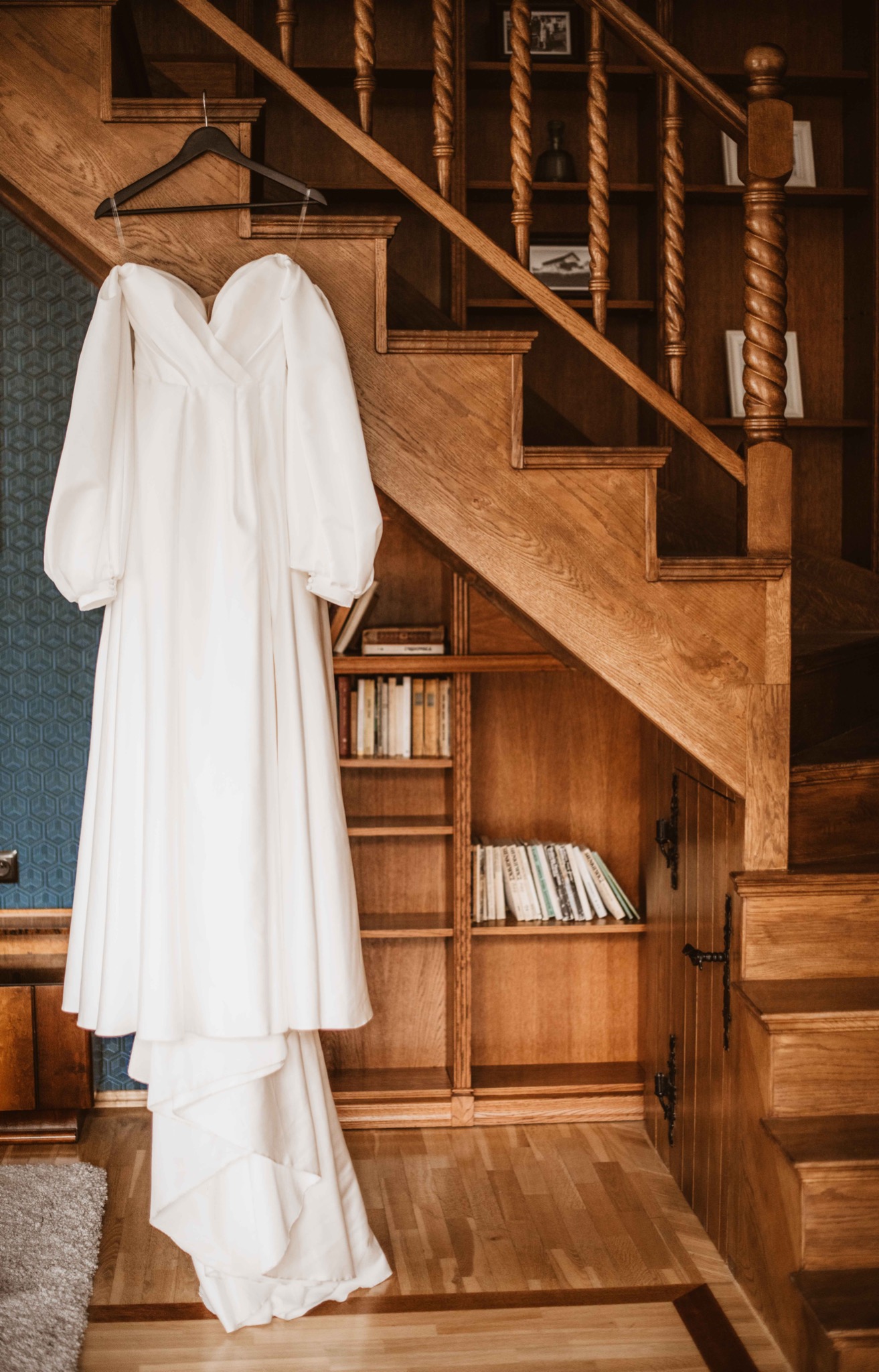 Elegancka suknia ślubna z bufiastymi rękawami, wisząca na wieszaku na tle drewnianych schodów i regałów z książkami.