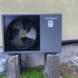 Szara pompa ciepła marki Nesheat zamontowana na betonowych bloczkach, z widocznym wentylatorem za kratką, rurą odpływową i fragmentem trawy.