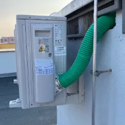 Zewnętrzna jednostka klimatyzacji marki Haier zamontowana na ścianie budynku z podłączonym zielonym, karbowanym przewodem wentylacyjnym, zabezpieczona metalową kratką